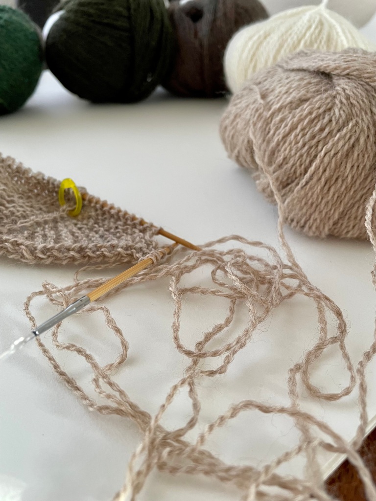 Taupe heather wool yarn against a white paper background, partially knit, partially jumbled out of a ball, with yarn in white and three balls forming a green gradient in the unfocused background 